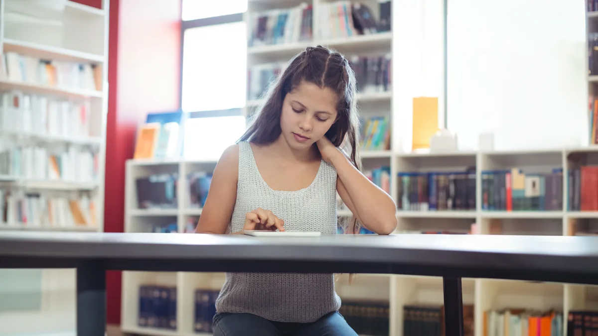 Estudiante concentrada usando una tableta en la biblioteca rodeada de estanterías de libros.