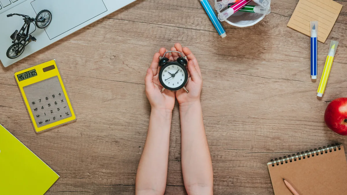 Manos sosteniendo un reloj despertador sobre un escritorio de estudio con calculadora y cuaderno.