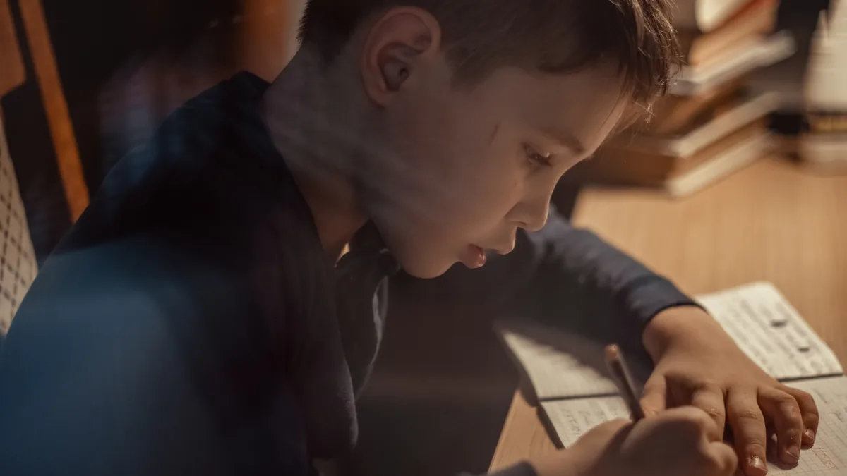 Niño concentrado escribiendo en su cuaderno mientras estudia junto a una pila de libros.