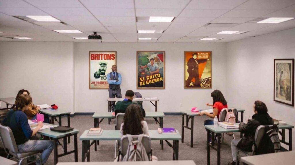 Profesor ante un grupo de estudiantes en un aula con carteles de propaganda en la pared.