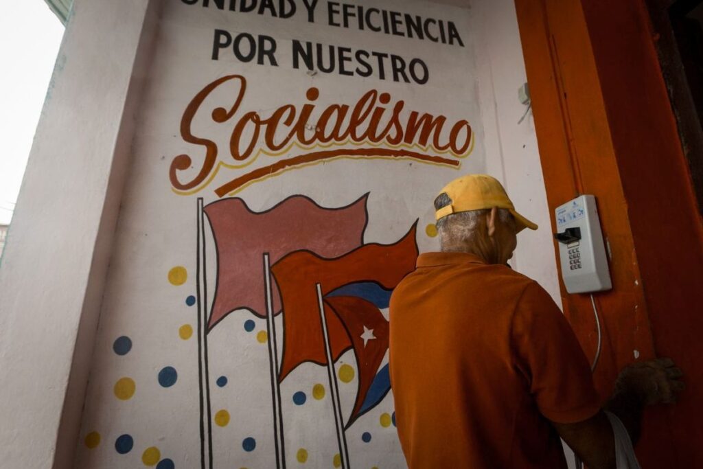 Hombre junto a un mural con banderas y el lema «Unidad y eficiencia por nuestro socialismo».